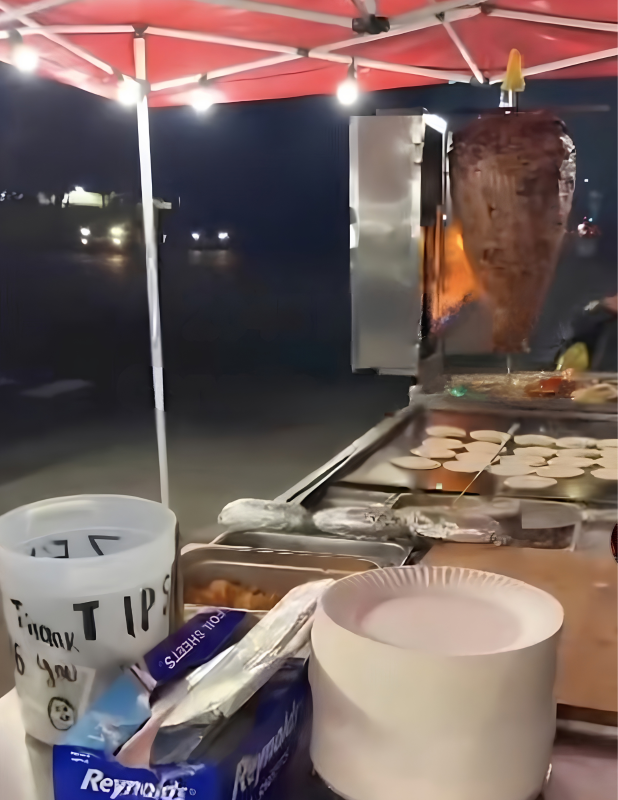 Meat is being cooked with an open flame under a pop-up tent. Other items are on a cook surface next to it, and some items appear to be finished and wrapped in foil on a makeshift counter. They are on a sidewalk at night, with headlights in the background.